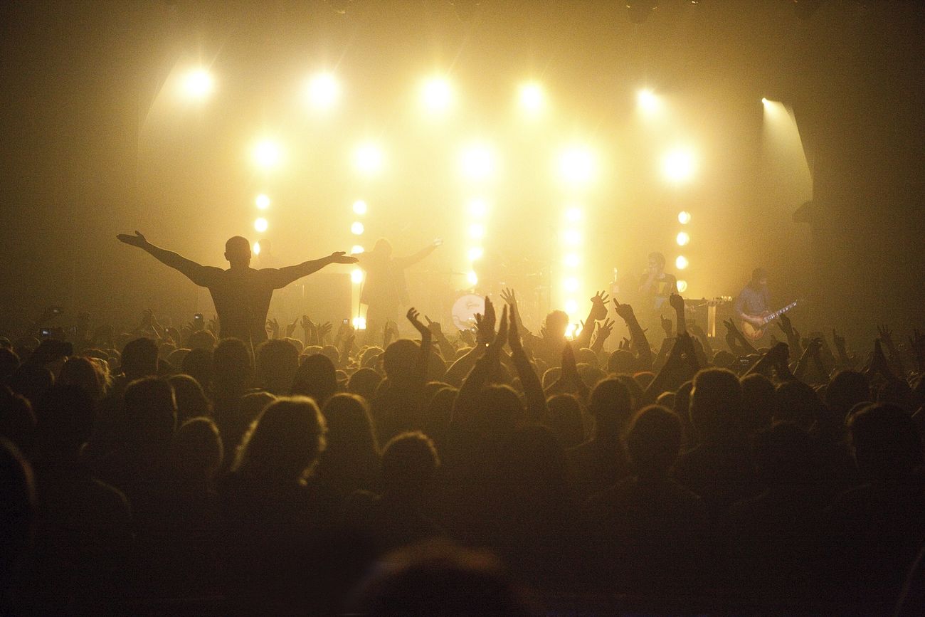 this is black and white image of a concert. the image is taken from the back of the room, with the stage at the centre of the image. In the foreground is the crowd, all of whom have their back to the camera, facing the stage. Lots of people in the crowd have their hands in the air and clapping. One person has been elevated from the crowd onto someone's shoulders, and has their arms outstretched. The stage has bright lights on it, meaning that the band members are hard to see