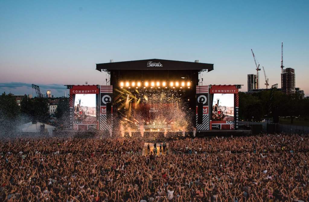 a black and white image of the the stage of finsbury park. The camera has taken the image from up high. in the centre of the image is the stage. There are lots of beams of lights coming from the stage, as well as confetti. In the bottom half of the image is the crowd with their back to the camera The top half of the image is the sky, with a few london sky scrapers on the right side, behind the stage. 