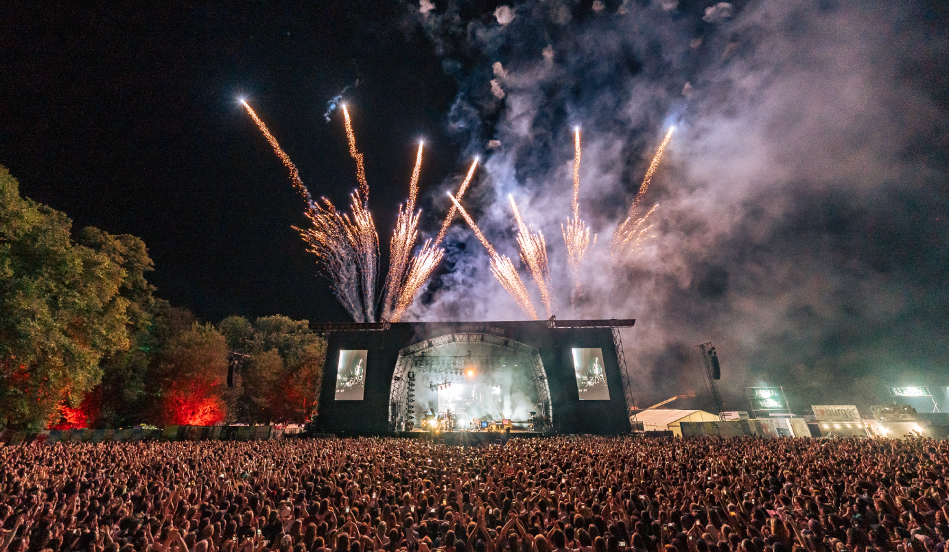 a black and white image of the stage from the middle of the crowd. The camera is up high and appears to have a mild fish eye lens. The stage is located in the middle of the bottom of the third of the stage. The stage is lit up by bright lights. Behind the stage is the night sky. There are trees on the left side in the top 3/4s of the image.  There are fireworks coming from the top of the stage, pointing upwards to the top of the image. The bottom 1/4 of the picture is of the crowd, all of whom have their backs facing the camera, facing the stage. There are thousands of people in the crowd.