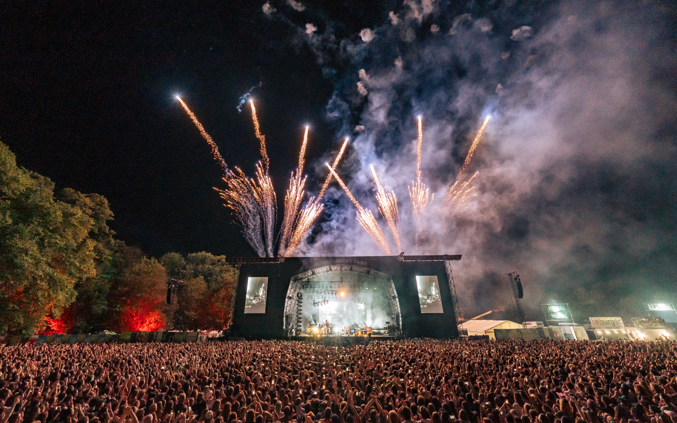 a black and white image of the stage from the middle of the crowd. The camera is up high and appears to have a mild fish eye lens. The stage is located in the middle of the bottom of the third of the stage. The stage is lit up by bright lights. Behind the stage is the night sky. There are trees on the left side in the top 3/4s of the image. There are fireworks coming from the top of the stage, pointing upwards to the top of the image. The bottom 1/4 of the picture is of the crowd, all of whom have their backs facing the camera, facing the stage. There are thousands of people in the crowd.