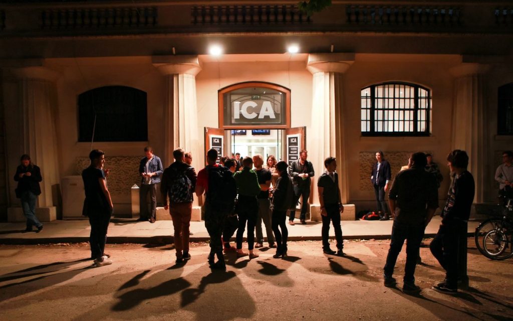 this is a black and white image of the entrace of the ICA. In the centre of the image is an open door, with large letters of ICA above it. There are two big pillars on either side of the door.The ICA is a stone, old looking building. The building takes up the top 2/3 of the image. The bottom 1/3 is of the pavement and the road. standing on the road and pavement are around 20 people. Most of them are chatting in small groups. It is at night time
