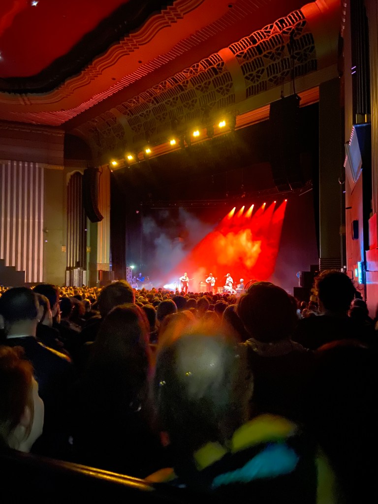 a colour image of the stage from the disability platform. the bottom half of the image is the crowd, all of whom have their backs to the camera facing the stage. in the centre of the top 1/3 of the image is the stage. it is lit up from behind by a row of red lights. the lights illuminate three figures at the front of the stage. in the middle is adrianne lenker holding a guitar. she is a white woman in her thirties with short dark hair. on either side of her are two white men with dark hair, both holding guitars. the camera is to the right of the stage