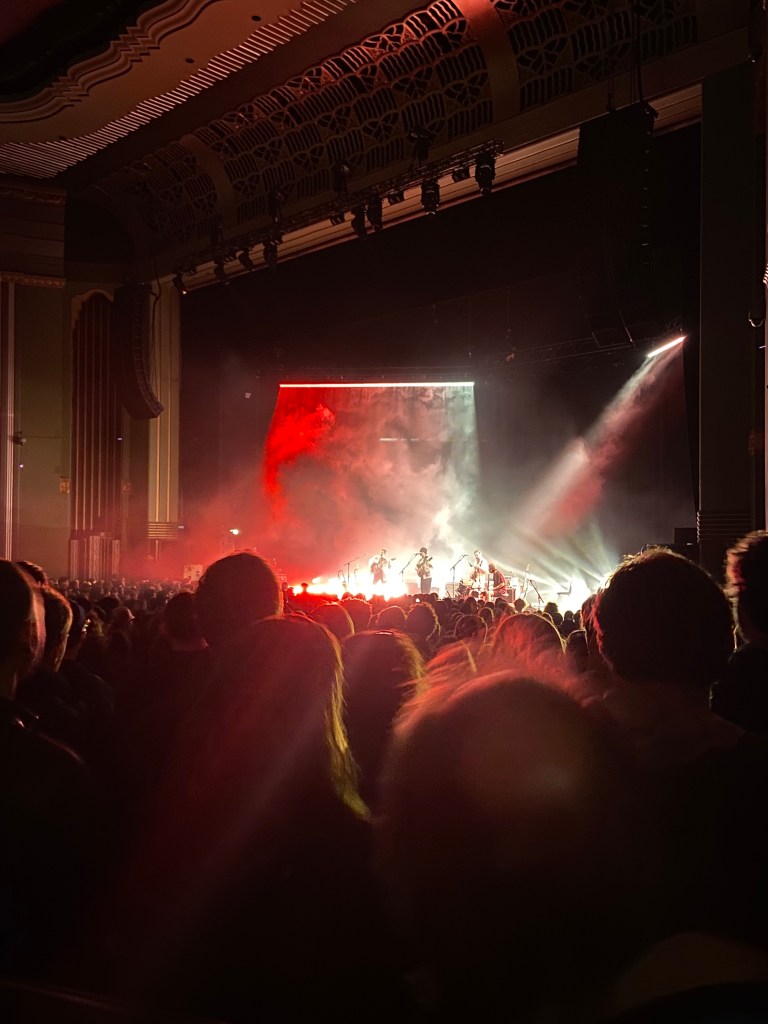 a colour image of the stage from the disability platform. the bottom half of the image is the crowd, all of whom have their backs to the camera facing the stage. in the centre of the top 1/3 of the image is the stage. it is lit up from behind by red, green and white lights. the lights illuminate three figures at the front of the stage. in the middle is adrianne lenker holding a guitar. she is a white woman in her thirties with short dark hair. on either side of her are two white men with dark hair, both holding guitars. the camera is to the right of the stage