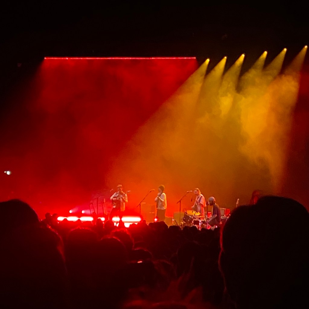 a colour image of the stage from the disability platform. the bottom 1/4 of the image is the crowd, all of whom have their backs to the camera facing the stage. in the centre of the image is the stage. it is lit up by red and yellow lights. there is a lot of red and yellow smoke. there are 4 figures on stage . in the middle is adrianne lenker holding a guitar and singing to a microphone. she is a white woman in her thirties with short dark hair. tp her left is a middle aged man with dark hair playing a guitar. on her right are two men, both with dark hair, on playing the piano and one holding a guitar. the camera is to the right of the stage