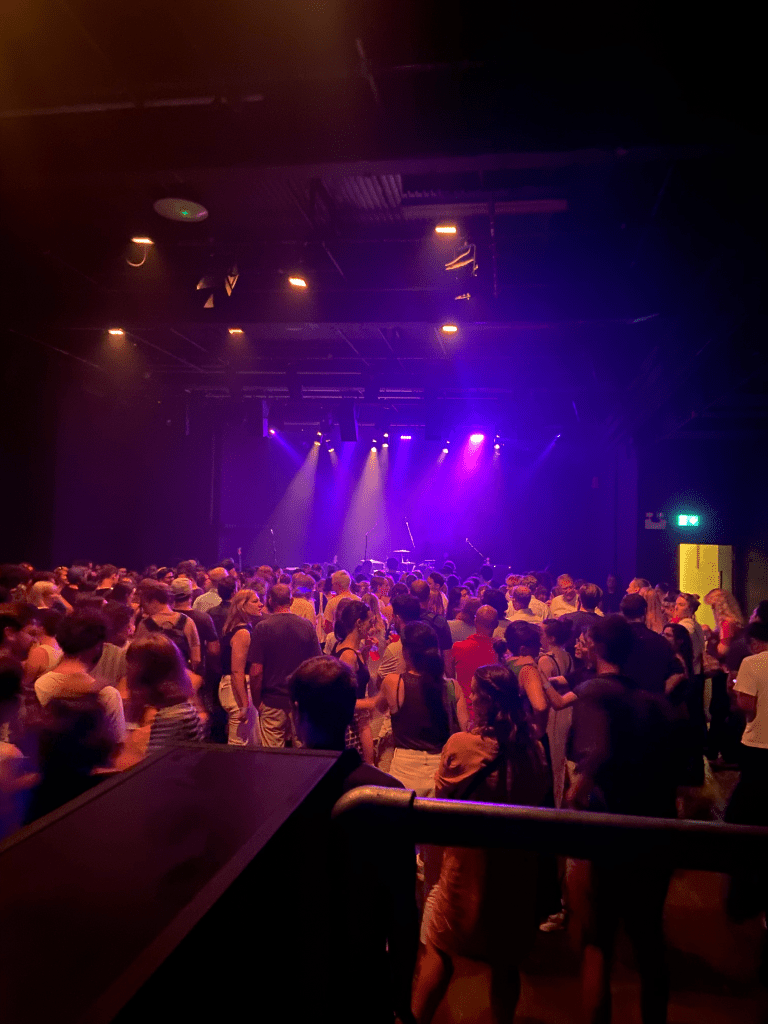 a colour image of the stage at the ICA from the back of the crowd. In the foreground on the image you can see the rail at the front of the platform. The stage is in the centre of the iamge at the back, and is lit up with purple lights. the stage is empty. it is a black room with black walls and ceiling. there are white lights on the ceiling. he bottom half of the image is of the crowd with their backs to the camera, facing the stage. there are a couple hundred people in the crowd