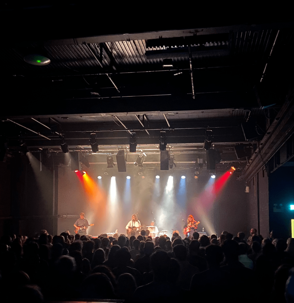 a colour image of the stage at the ICA from the back of the crowd. in the foreground on the image you can see the rail at the front of the platform. the stage is in the centre of the image at the back, and is lit up with red white and blue lights. the stage has 4 people on it. marika hackman is a white woman holding a guitair in the middle of the stage, wearing a white shirt. she has short brown hair. to the left of her is a man holding a guitar. to the right of her is a woman with long hair holding a guitar. behind the three of them is a woman with short dark hair playing the drums. it is a black room with black walls and ceiling. the bottom half of the image is of the crowd with their backs to the camera, facing the stage. there are a couple hundred people in the crowd