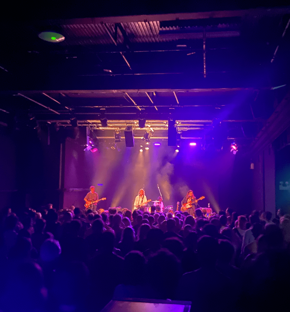 a colour image of the stage at the ICA from the back of the crowd. in the foreground on the image you can see the rail at the front of the platform. the stage is in the centre of the image at the back, and is lit up with purple lights. the stage has 4 people on it. marika hackman is a white woman holding a guitar in the middle of the stage, wearing a white shirt. she has short brown hair. to the left of her is a man holding a guitar. to the right of her is a woman with long hair holding a guitar. behind the three of them is a woman with short dark hair playing the drums. it is a black room with black walls and ceiling. the bottom half of the image is of the crowd with their backs to the camera, facing the stage. there are a couple hundred people in the crowd