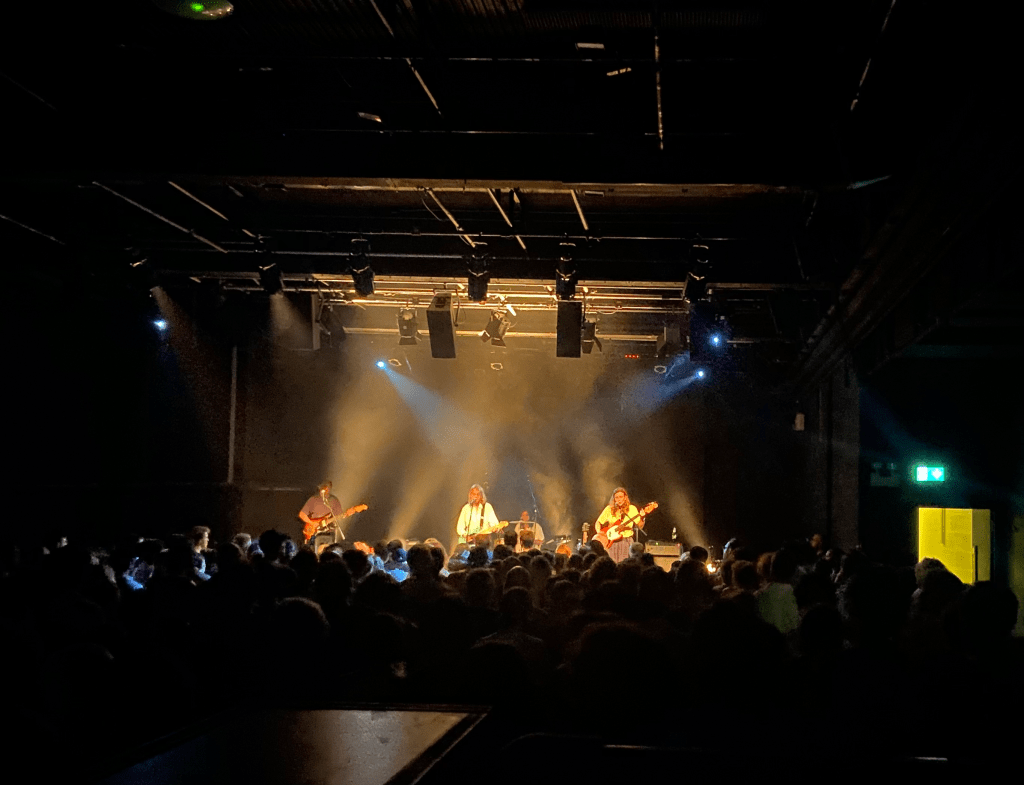 a colour image of the stage at the ICA from the back of the crowd. in the foreground on the image you can see the rail at the front of the platform. the stage is in the centre of the image at the back, and is lit up with yellow lights. the stage ishas 4 people on it. marika hackman is a white woman holding a guitair in the middle of the stage, wearing a white shirt. she has short brown hair. to the left of her is a man holding a guitar. to the right of her is a woman with long hair holding a guitar. behind the three of them is a woman with short dark hair playing the drums. it is a black room with black walls and ceiling. the bottom half of the image is of the crowd with their backs to the camera, facing the stage. there are a couple hundred people in the crowd