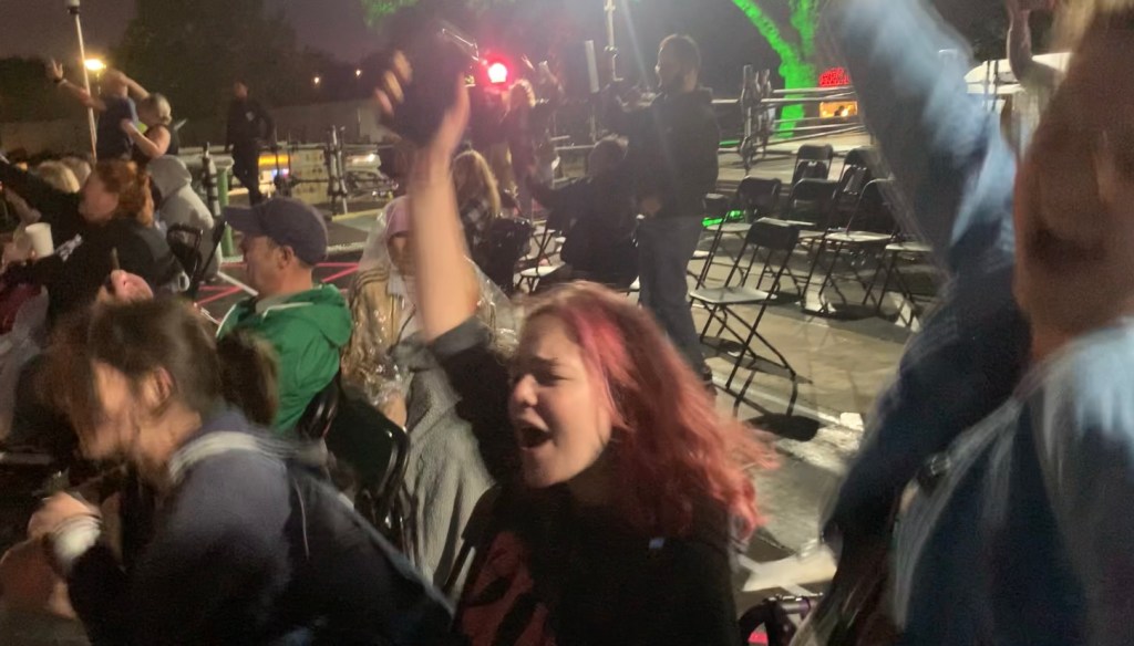 a black and white image of the disability platform. in the centre of the image is a white, 21 year old woman. she is sat in a wheelchair.. she has her eyebrow pierced. she is has an arm up and is singing along to the music. she is full of joy. . to the right of her are a line of people sat down watching the gig. some have their hands up, some have phones out recording. behind the line of people is the rest of the platform, mainly empty with a few individual people stood up scattered across the platform. there are about 15-20 people in the image total. it is taking place at night time