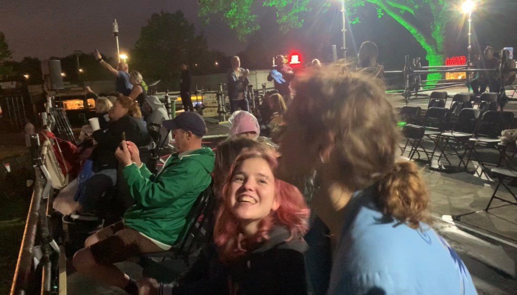 a black and white image of the disability platform. in the centre of the image is a white, 21 year old woman. she has her eyebrow pierced. she is sat in the wheelchair she is looking up at her friend to the left of her, with a big grin on her face. to the right of her are a line of people sat down watching the gig. some have their hands up, some have phones out recording. behind the line of people is the rest of the platform, mainly empty with a few individual people stood up scattered across the platform. there are about 15-20 people in the image total. it is taking place at night time
