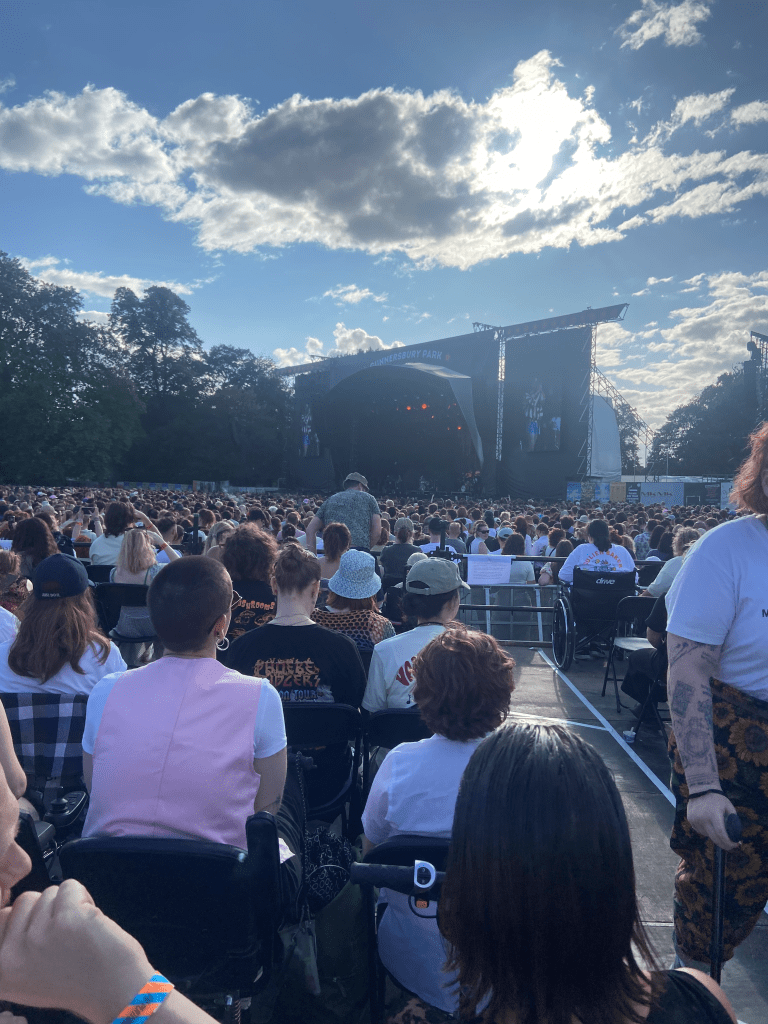 a colour image of the stage from the platform. the bottom 2/3s of the image is of the crowd, all of whom have their backs to the camera facing the stage. due to the angle, much of the picture shows people sat in the disabled platform. there are around 100 people or so on the platform, sat in rows. the stage is far away and hard to see. the stage is dark. behind the stage is a blue sky with some clouds. the stage and sky take up the top 1/3 of the picture