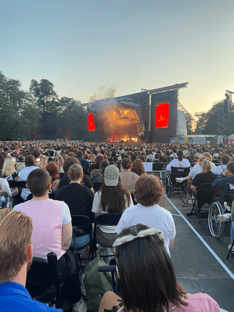 a colour image of the stage from the platform. the bottom 2/3s of the image is of the crowd, all of whom have their backs to the camera facing the stage. due to the angle, much of the picture shows people sat in the disabled platform. there are around 100 people or so on the platform, sat in rows. the stage is far away and hard to see. the stage is lit up by red and orange light. there are some figures perhaps playing musical instruments. there is smoke on the stage. on either side of the stage are screens lit up red. behind the stage is a blue sky with some clouds. the stage and sky take up the top 1/3 of the picture