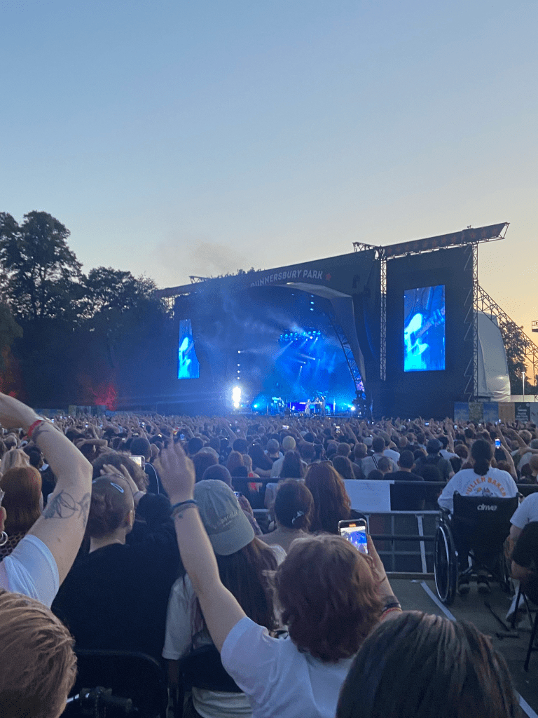 a colour image of the stage from the platform. the bottom 2/3s of the image is of the crowd, all of whom have their backs to the camera facing the stage. due to the angle, much of the picture shows people sat in the disabled platform. there are around 100 people or so on the platform, sat in rows. a few of the people have their arms up, swaying to the music. the stage is far away and hard to see. the stage is lit up by blue lights. there are some figures perhaps playing musical instruments. there is smoke on the stage. on either side of the stage are screens lit up blue. behind the stage is a blue and yellow sky, likely just after sunset. the stage and sky take up the top 1/3 of the picture