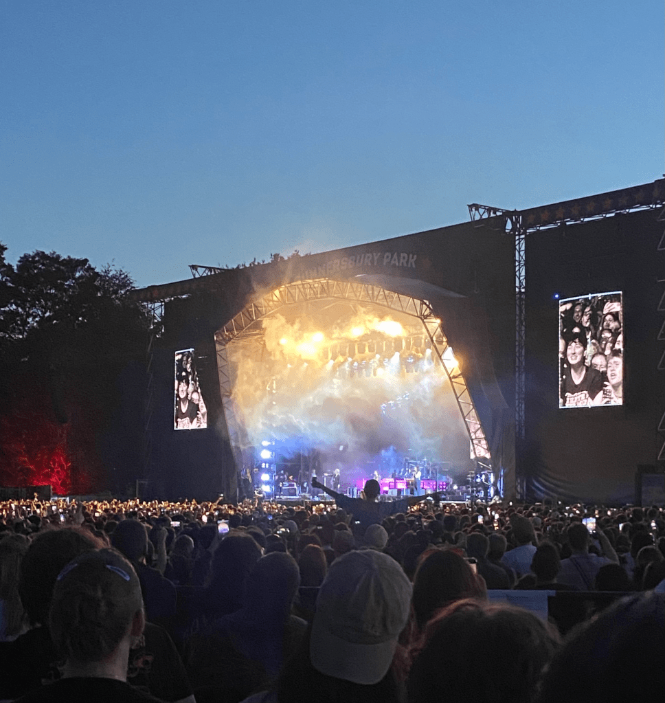 a colour image of the stage from the platform. the bottom 1/3 of the image is of the crowd, all of whom have their backs to the camera facing the stage.  the stage is far away and hard to see. the stage is lit up by a gradient of lights, going from yellow to blue to purple. there are some figures perhaps playing musical instruments. there is smoke on the stage. on either side of the stage are screens, showing black and white images of fans watching the show. one person is above the crowd, most likely on someone's shoulders, with their arms outstretched. behind the stage is a dark blue sky - it is dust. the stage and sky take up the top 2/3 of the picture