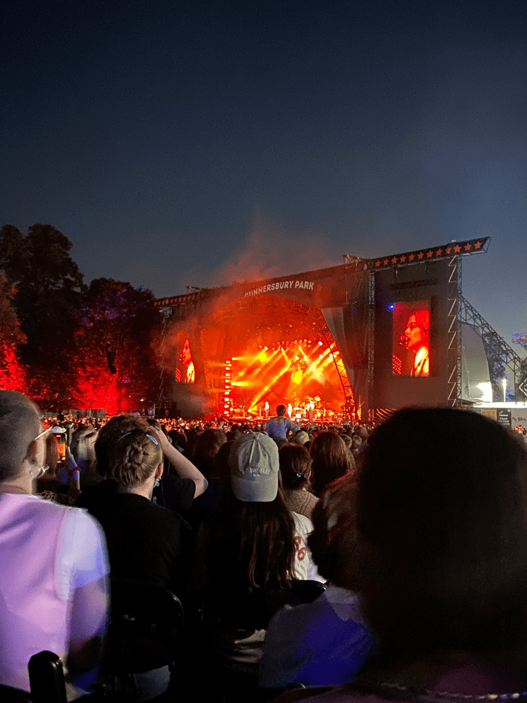 a colour image of the stage from the platform. the bottom 2/3s of the image is of the crowd, all of whom have their backs to the camera facing the stage. due to the angle, much of the picture shows people sat in the disabled platform. there are around 100 people or so on the platform, sat in rows. the stage is far away and hard to see. the stage is lit up by red and orange light. there are some figures perhaps playing musical instruments. there is smoke on the stage. on either side of the stage are screens lit up red with an image of a young white woman singing to a microphone. behind the stage is a dark blue sky, it is dusk. the stage and sky take up the top 1/2 of the picture.
