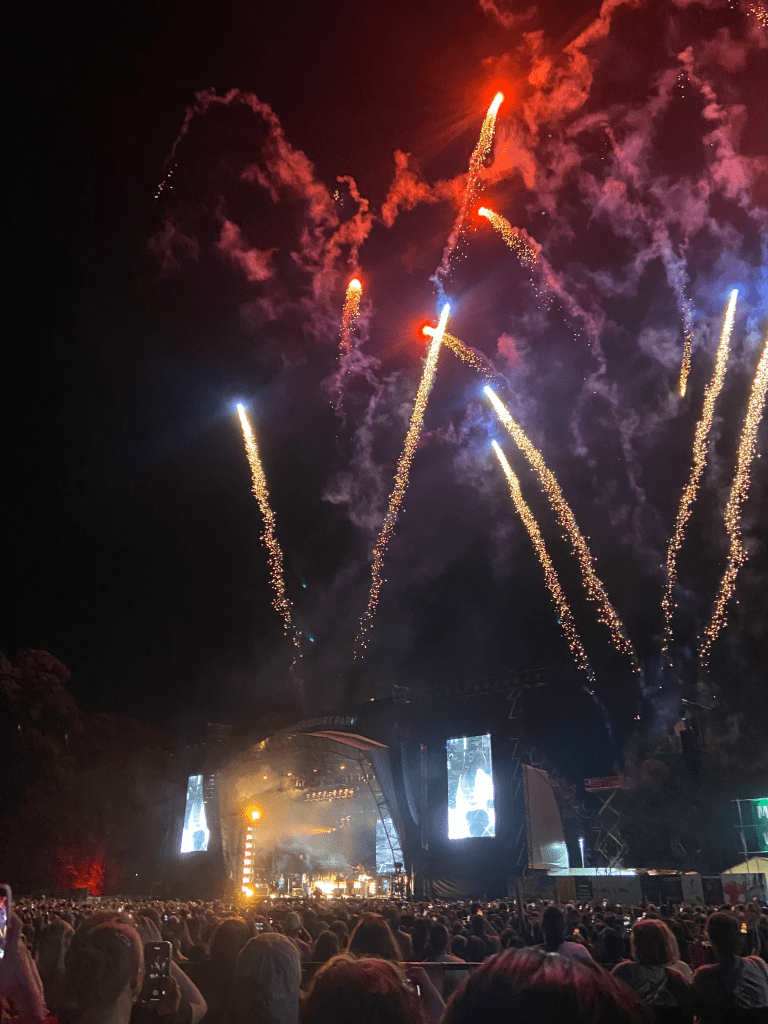 a colour image of the stage from the platform. the bottom 1/4s of the image is of the crowd and the stage, while the rest of the image is of fireworks coming from the stage, with the night sky in the background. the fireworks are red, blue and yellow. the stage is in the bottom left corner. it is hard to see. it is lit up by yellow lights and smoke. on either side of the stage are screens lit up in blue, showing close ups of the stage. below the stage shows the crowd, all of whom have their backs to the camera facing the stage. the stage is far away and hard to see. there are some figures perhaps playing musical instruments.