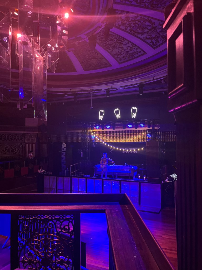 An image taken from the accessible platform platform. At the bottom of the image you can see the bars of the platform. In the centre of the image is the stage with a sofa on it. There is a person on the stage, in front of the sofa, speaking into a microphone. Above the sofa is two strings of fairy lights and three lit up drawings of a tooth. The room is lit up with red lighting