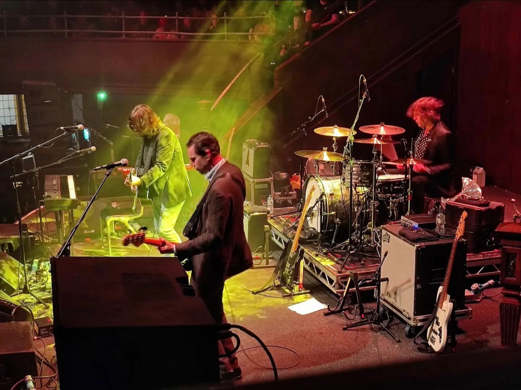the image shows a close up view of the band. the person taking the photo is a few feet away from them, to hte left. in the picture, a white man in the foreground plays guitar, behind him another white man with long hair also plays guitar. behind them and to the right of the image another band member plays the drums. there is a green spotlight that illuminates the band.